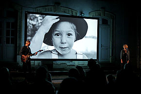 Sur une scène, un guitariste et une comédienne devant un micro. Derrière eux, un écran avec une petite fille qui porte un chapeau et une salopette. Devant eux le public. 