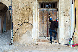 Un homme avec un blouson noir et un bonnet rouge, blanc et bleu est face à une porte en bois. Il tient une perche de prise de son. - Agrandir l'image (fenêtre modale)