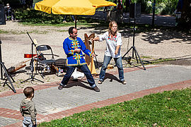 Une femme et un homme réalisent un spectacle dans un parc. Il y a des instruments de musique : harpe et guitare. Un enfant se trouve au premier plan.