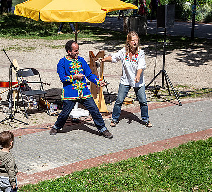 Une femme et un homme réalisent un spectacle dans un parc. Il y a des instruments de musique : harpe et guitare. Un enfant se trouve au premier plan.