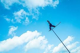 À plusieurs mètres de hauteur, une femme, habillée tout en bleu, se tient debout sur une tige bleue. En arrière fond, le ciel et les nuages. - Agrandir l'image (fenêtre modale)