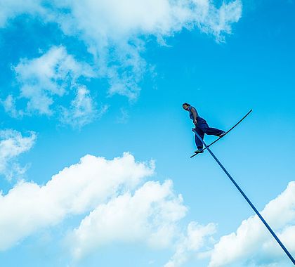 À plusieurs mètres de hauteur, une femme, habillée tout en bleu, se tient debout sur une tige bleue. En arrière fond, le ciel et les nuages.